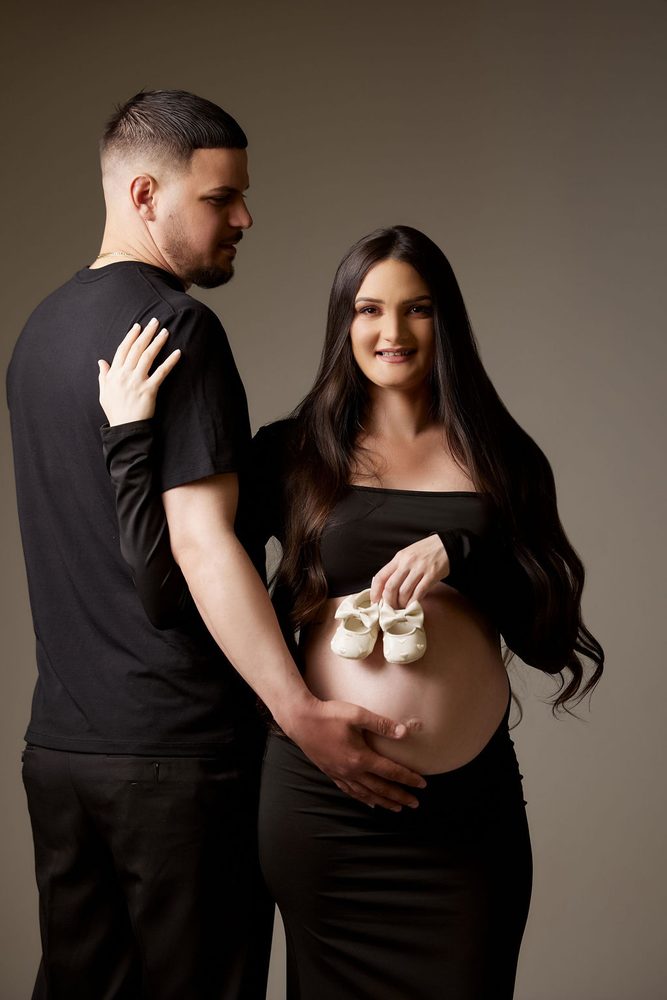 Expectant mother holding tiny white baby shoes against her belly as her partner stands beside her