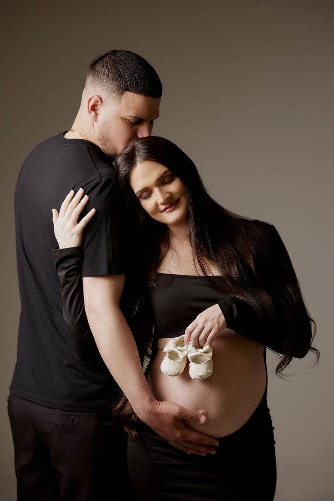 Partner kisses the top of the expectant mother's head as she holds bow-topped baby shoes over her bump