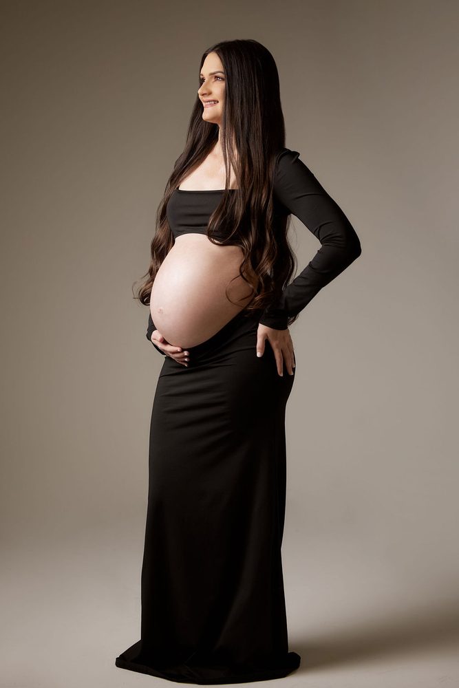 Full-length studio portrait of an expectant mother in a black crop top and maxi skirt, smiling upward