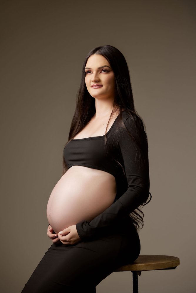 Expectant mother seated on a wooden stool, cradling her bare belly beneath a long-sleeve crop top