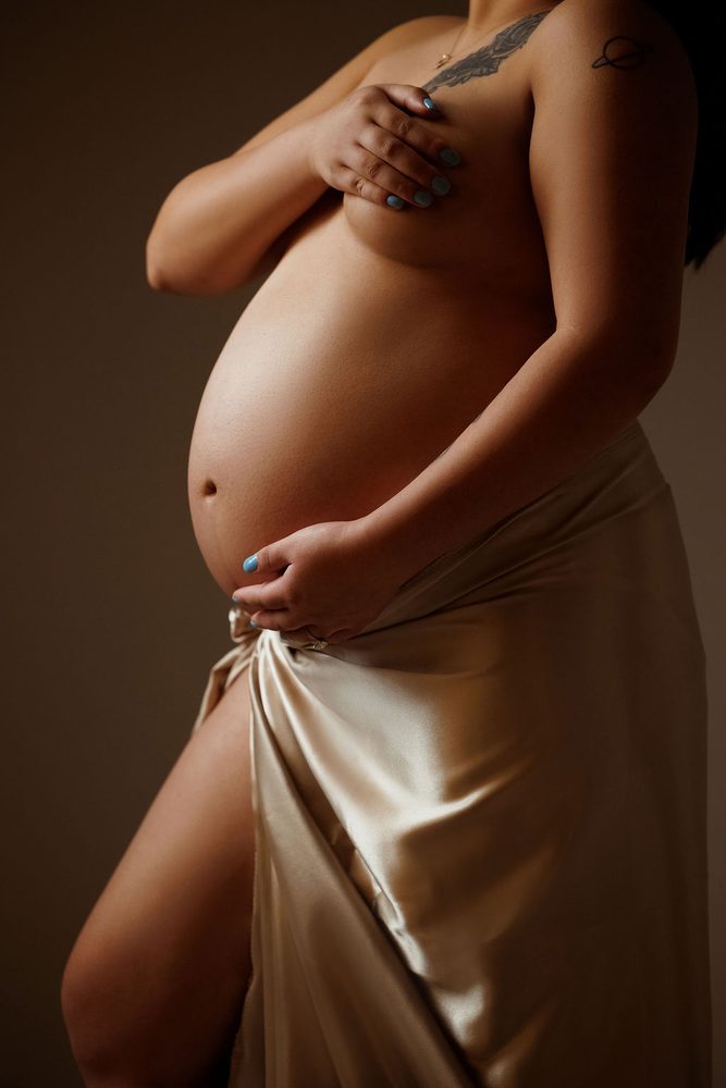 Close-up of a bare pregnant belly with a champagne satin drape at the hips in warm, low-key light