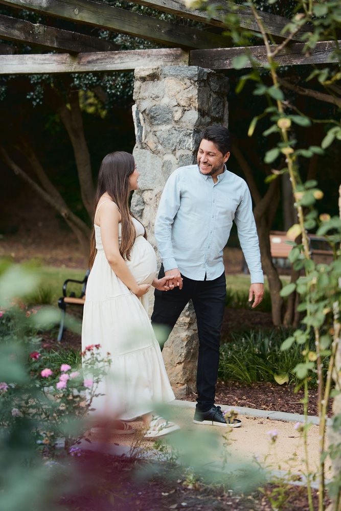 Expectant couple holding hands beneath a stone-pillared pergola in a rose garden