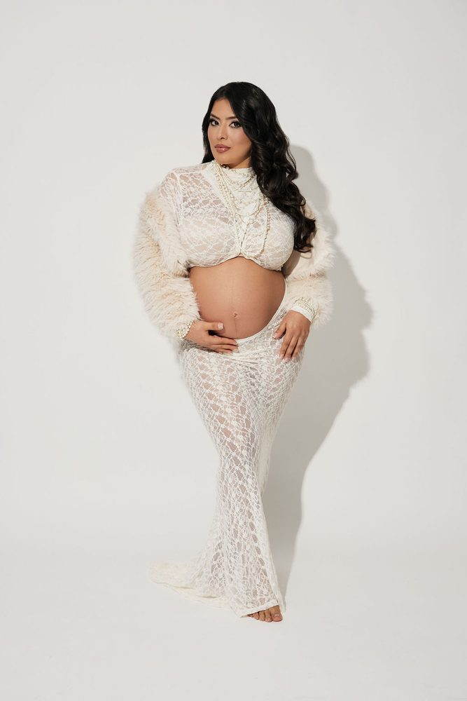Full-length portrait of an expectant mother in a white lace skirt set, faux-fur jacket and layered pearls