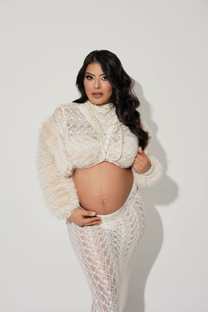 Expectant mother in a cream lace crop top and faux-fur shrug, pearls layered at her neck, hand on her bump