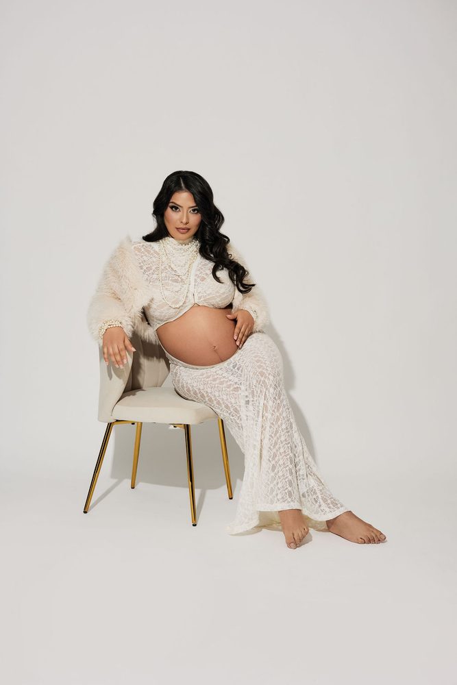 Expectant mother seated on a cream chair with gold legs, wearing white lace and a faux-fur jacket