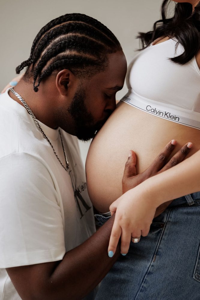 Partner in a white tee kisses the expectant mother's bare belly as she rests her hand on his