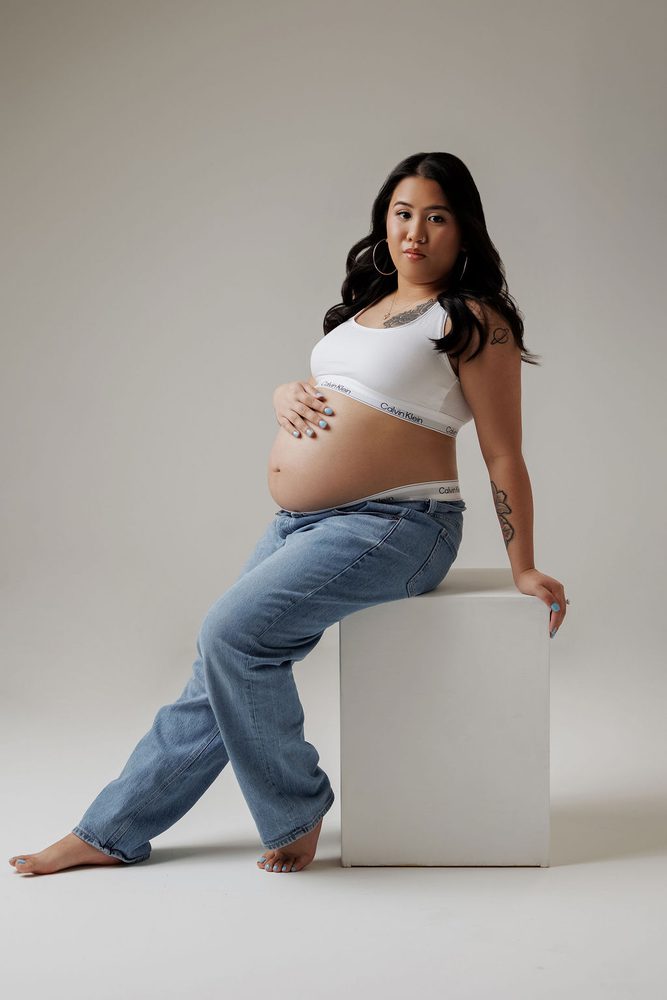 Expectant mother in a white sports bra and relaxed jeans sitting barefoot on a white studio cube