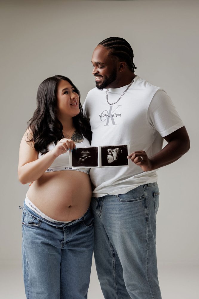 Expectant couple in white tees and jeans smiling at each other while holding up ultrasound prints