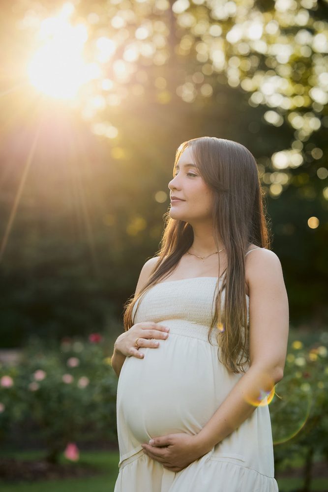 Expectant mother in profile at golden hour, sun flaring through the trees as she holds her belly