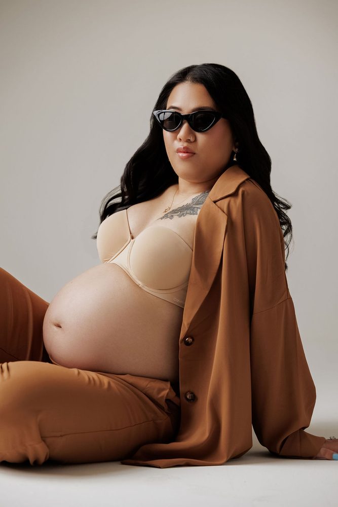 Expectant mother in cat-eye sunglasses and a camel shirt and trousers reclining on the studio floor