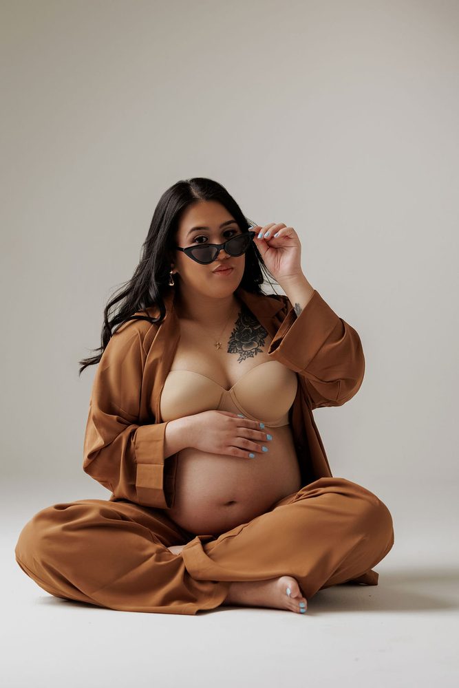 Expectant mother sitting cross-legged in a camel lounge set, lowering her cat-eye sunglasses