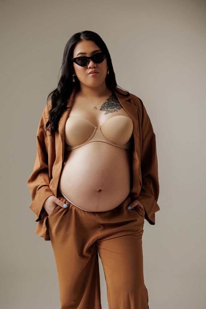 Expectant mother standing with hands in her pockets, open camel shirt and cat-eye sunglasses