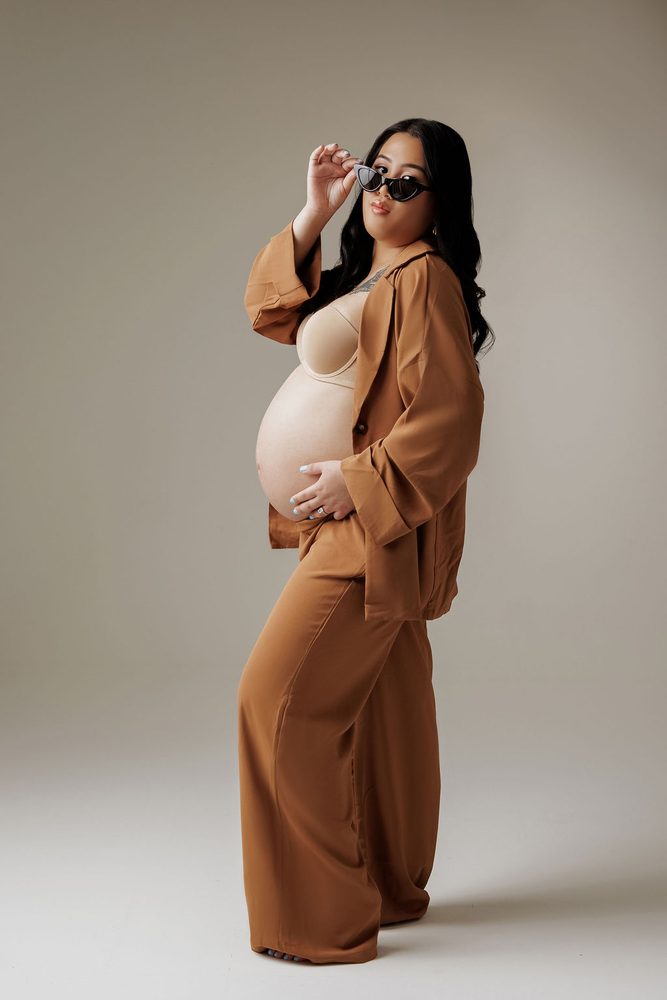 Full-length side pose in a caramel wide-leg set, tipping her sunglasses with one hand on her bump