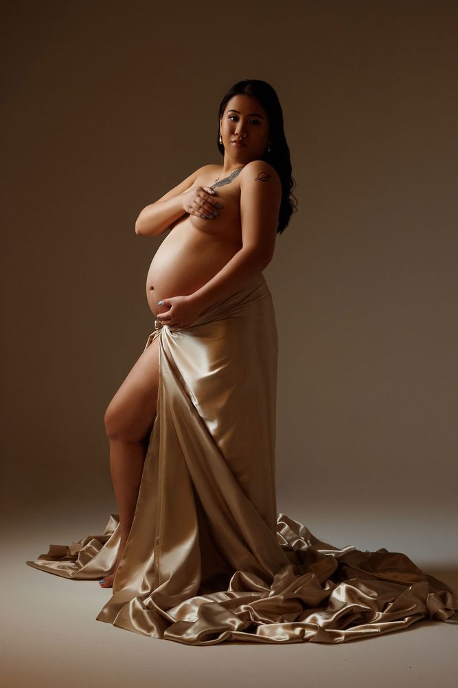 Full-length portrait of an expectant mother wrapped in flowing champagne satin under dramatic studio light