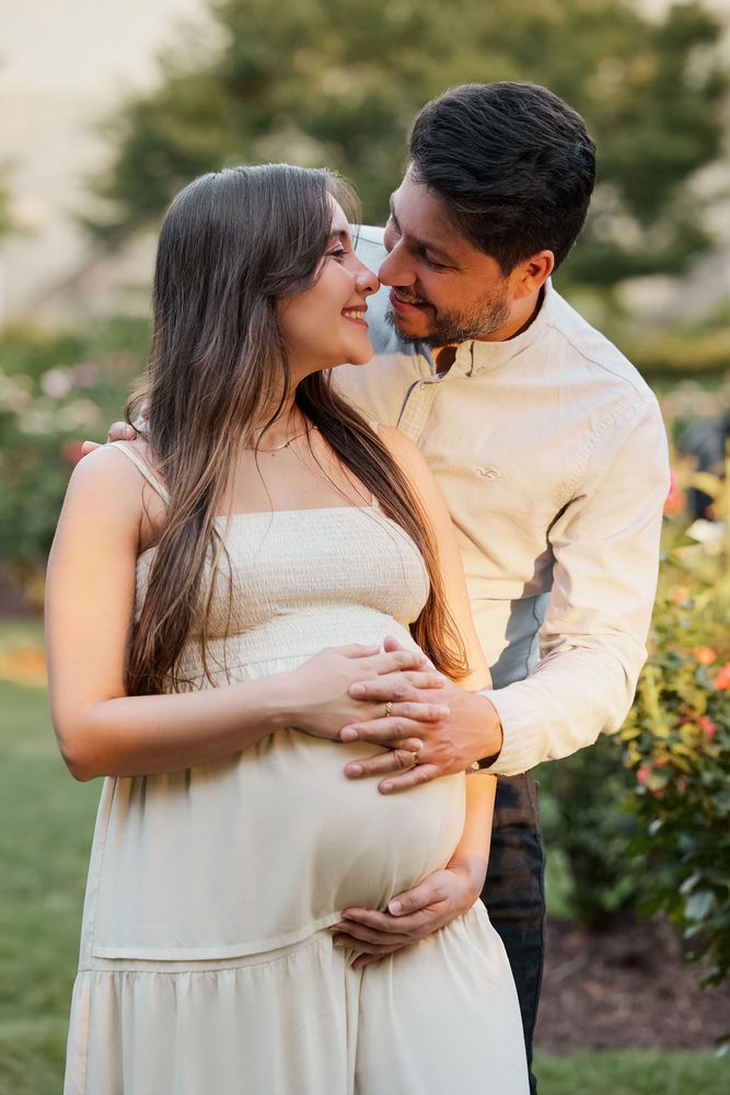 Expectant couple nose to nose in a garden, his hands layered over hers on her belly
