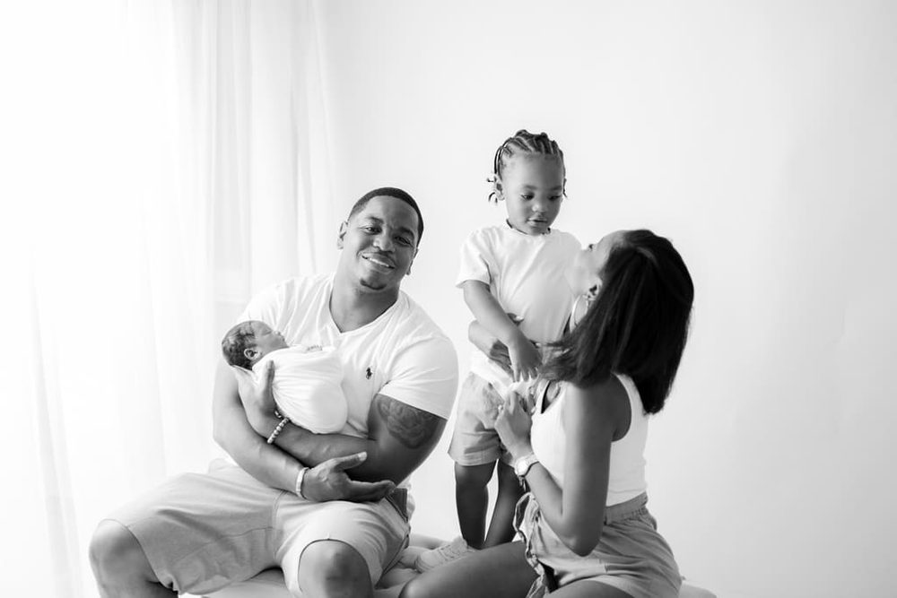 Black-and-white family portrait, smiling father cradling a swaddled newborn as mother plays with their toddler son