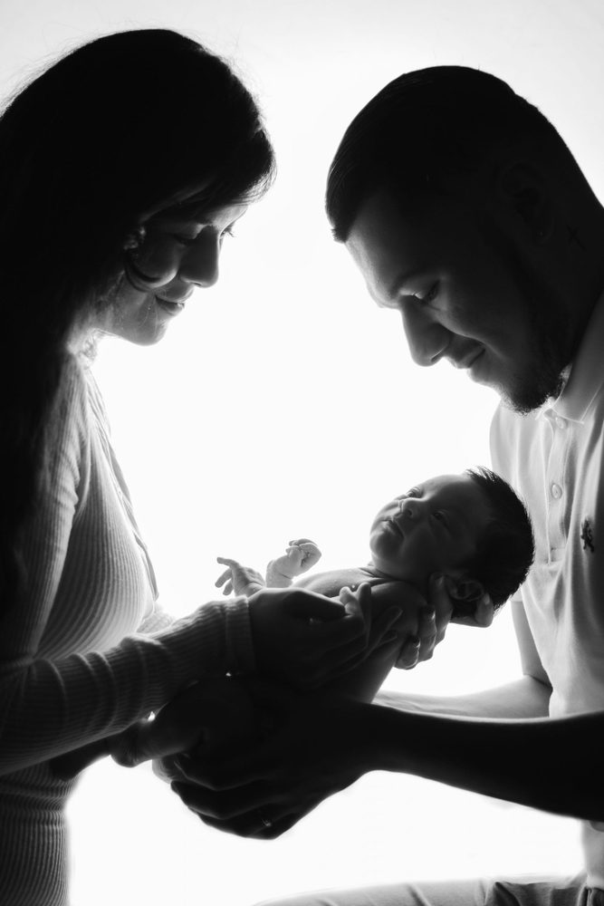 Backlit black-and-white profile of mother and father smiling down at the newborn held between their hands