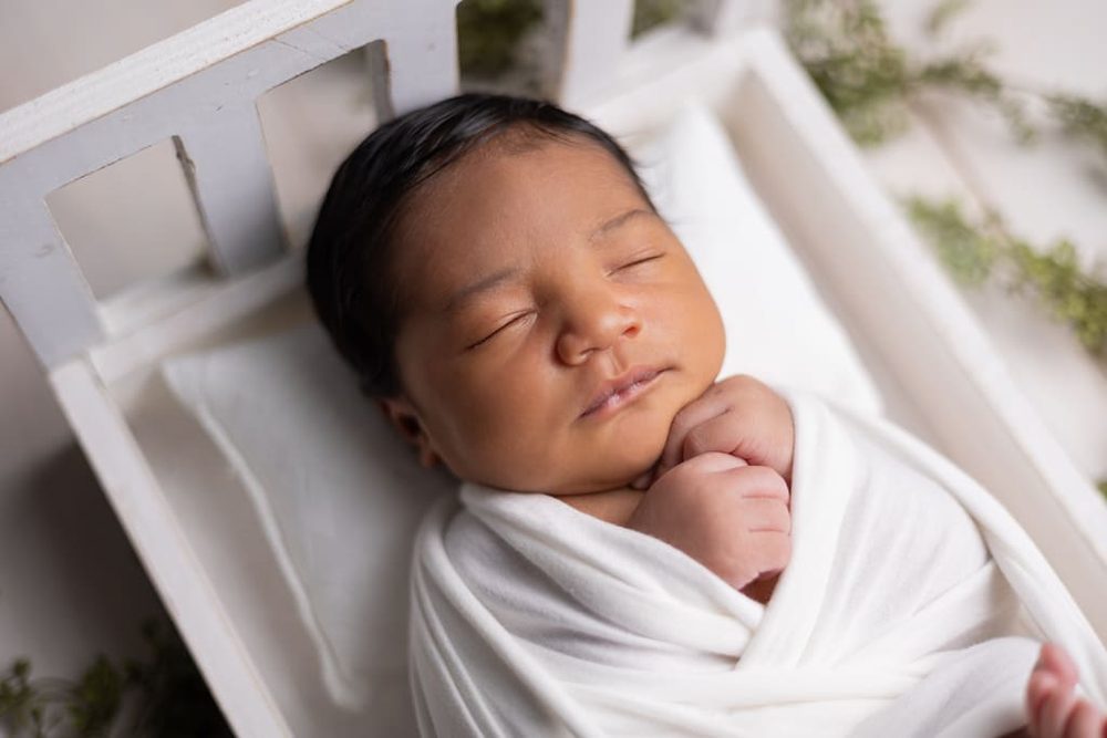 Sleeping newborn swaddled in white with hands under chin, resting in a small white wooden bed prop
