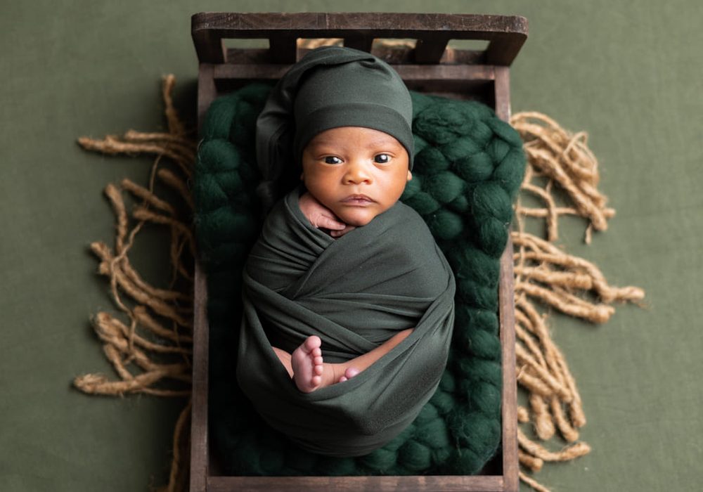 Alert newborn in a dark green swaddle and hat lying in a wooden bed on chunky green knit, jute fringe around