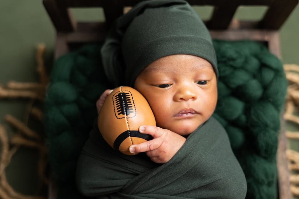 Newborn in a green hat and wrap holding a tiny leather football against his cheek