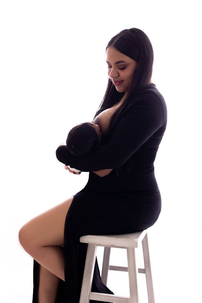 Mother in a black gown seated on a white stool breastfeeding her newborn against a bright white backdrop
