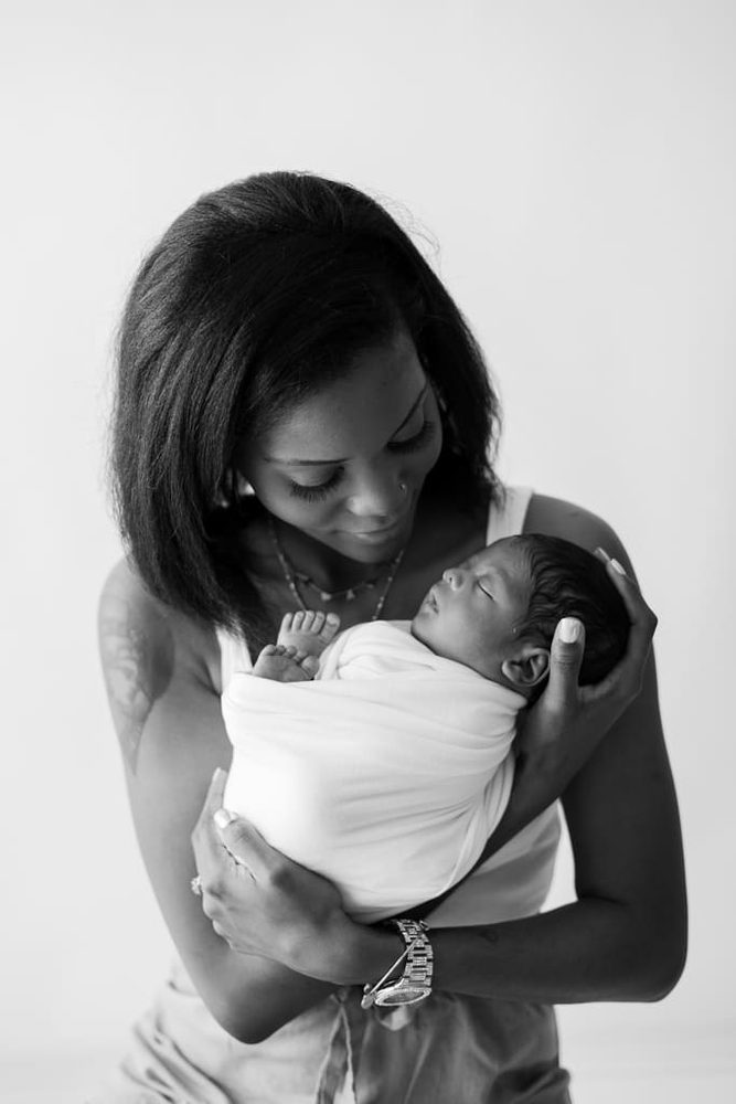 Black-and-white portrait of a mother gazing down at her sleeping newborn wrapped in white, held close to her chest
