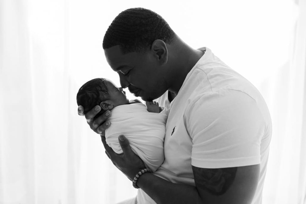 Black-and-white profile of a father touching noses with his swaddled newborn, backlit by a bright window