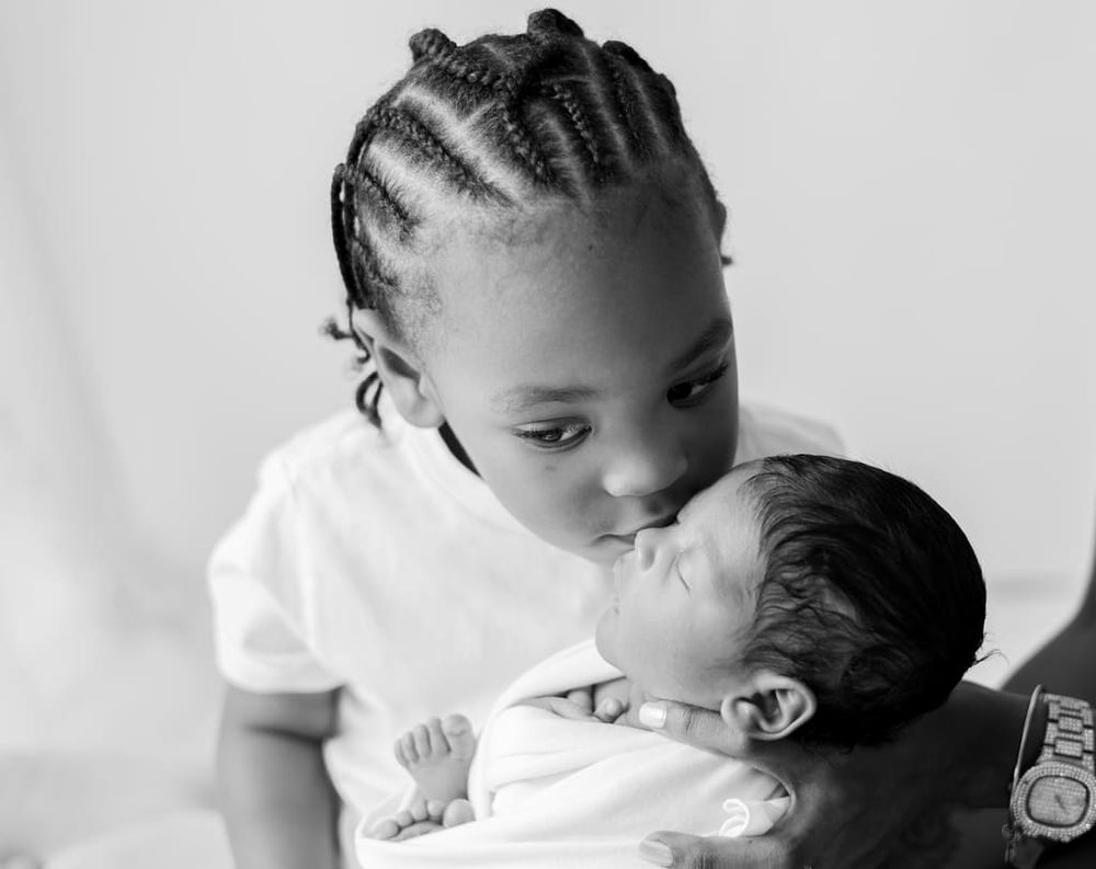 Black-and-white close-up of a big brother with braids kissing his sleeping newborn sibling's forehead