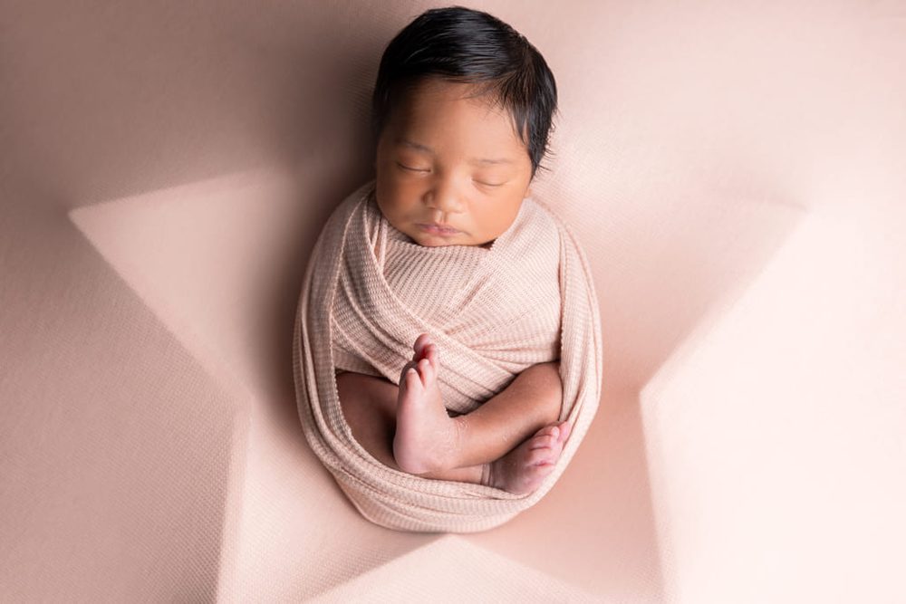 Sleeping newborn wrapped in a blush knit swaddle with tiny feet crossed, on a soft pink backdrop