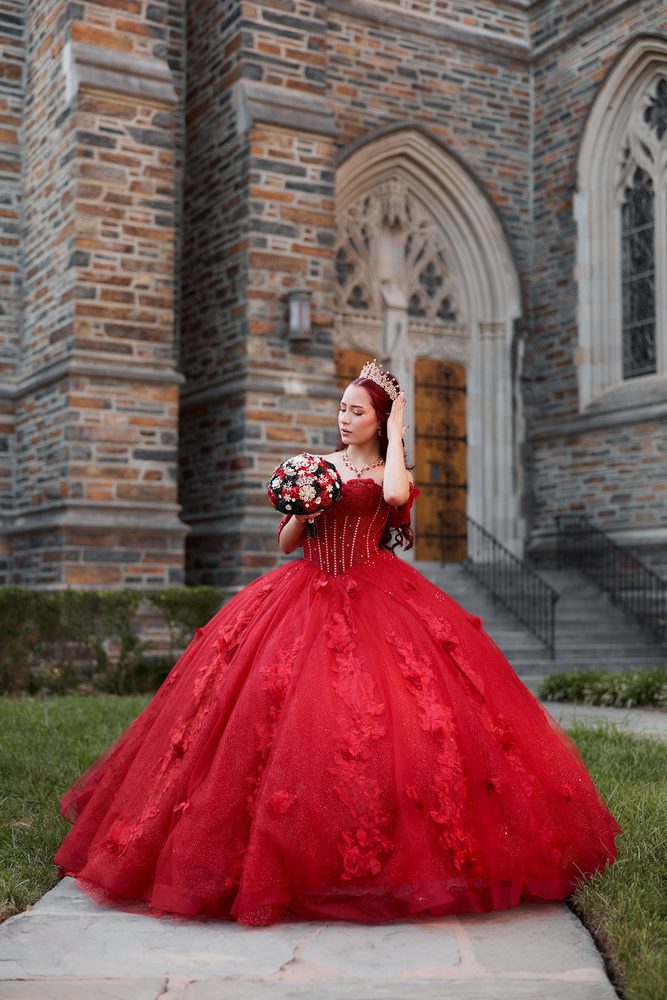 Quinceañera in a full red tulle gown with floral appliqués standing on a stone path by a Gothic chapel doorway