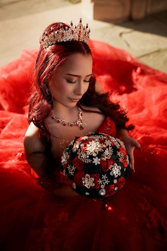 Overhead view of a quinceañera in a jeweled crown looking down at her red and black brooch bouquet, gown spread around her