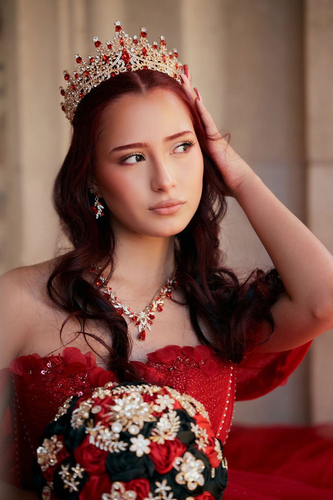 Close portrait of a quinceañera in a red-jeweled tiara and necklace, hand in her hair, beside a stone column