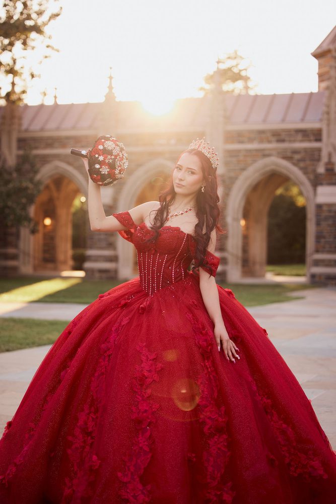 Quinceañera raising her bouquet overhead in a sunlit courtyard with stone archways and lens flare