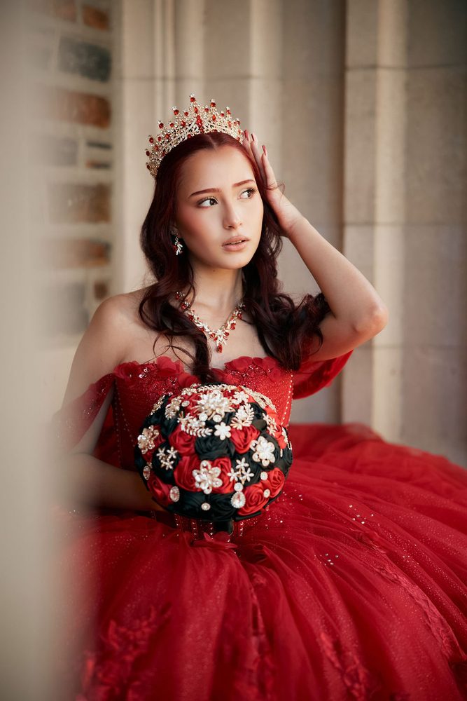 Seated quinceañera in a red off-the-shoulder gown and crown, bouquet in her lap, hand resting in her hair