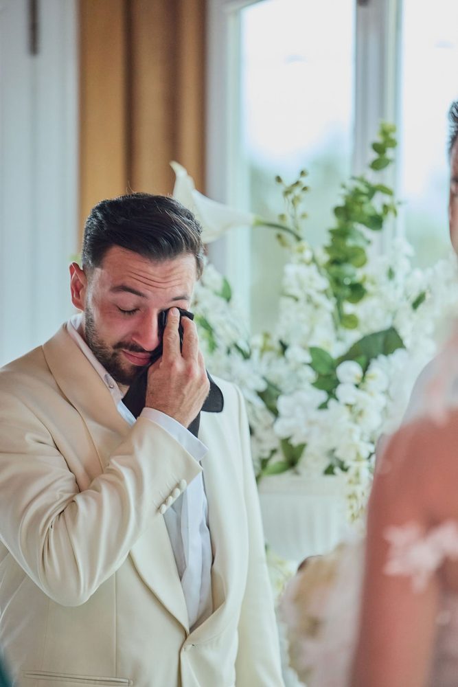 Groom in a cream dinner jacket wiping away tears with a handkerchief as his bride approaches