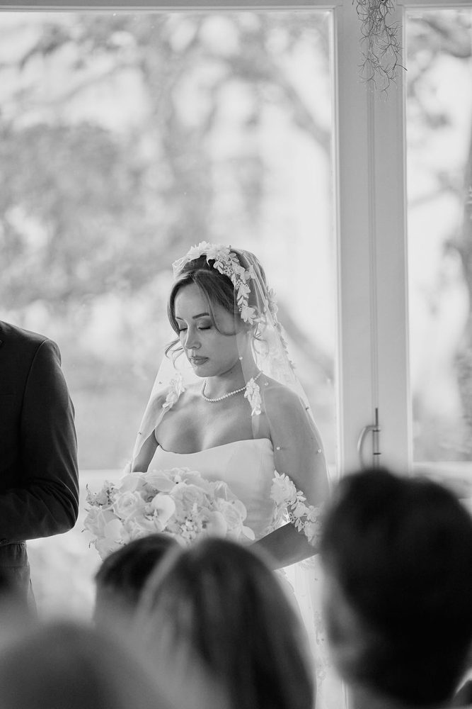 Black-and-white portrait of a bride with eyes closed during the ceremony, floral lace veil, pearls and rose bouquet