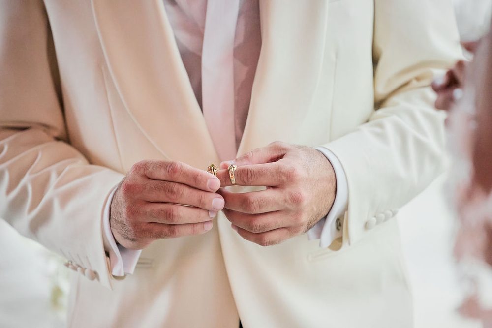 Close-up of the groom's hands holding two gold wedding bands against his cream tuxedo jacket