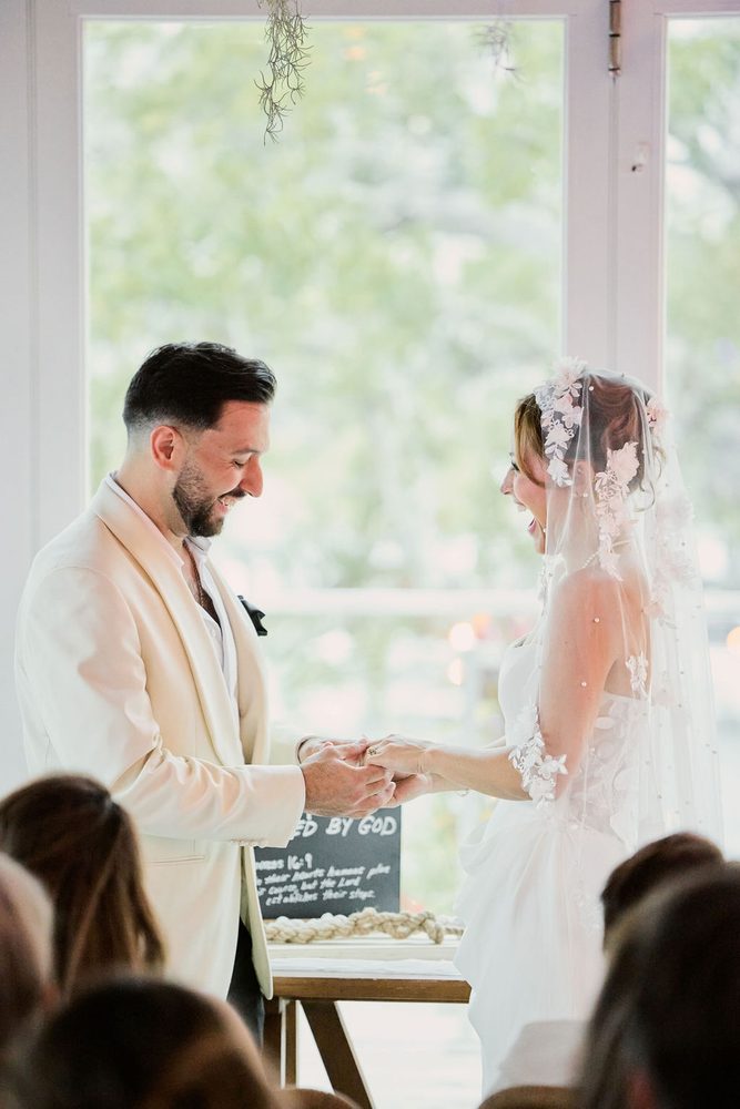Smiling bride and groom exchanging rings before a tall window at an indoor ceremony, guests in the foreground
