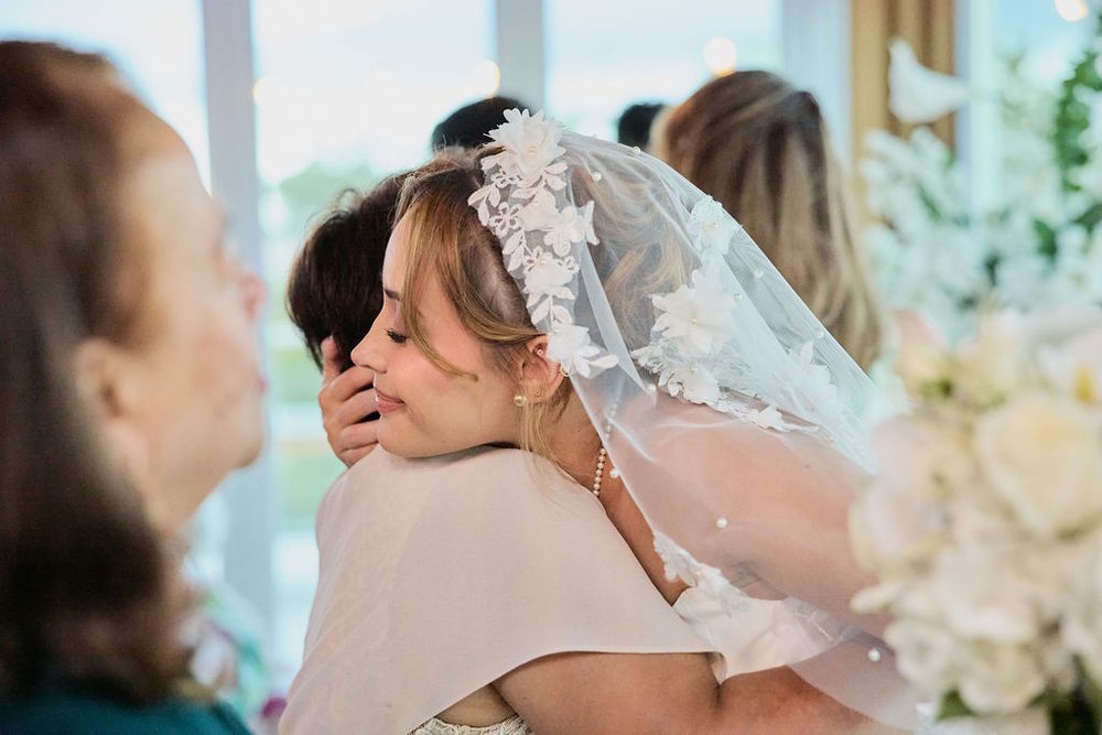 Bride in a flower-trimmed lace veil sharing a tearful hug with a guest after the ceremony