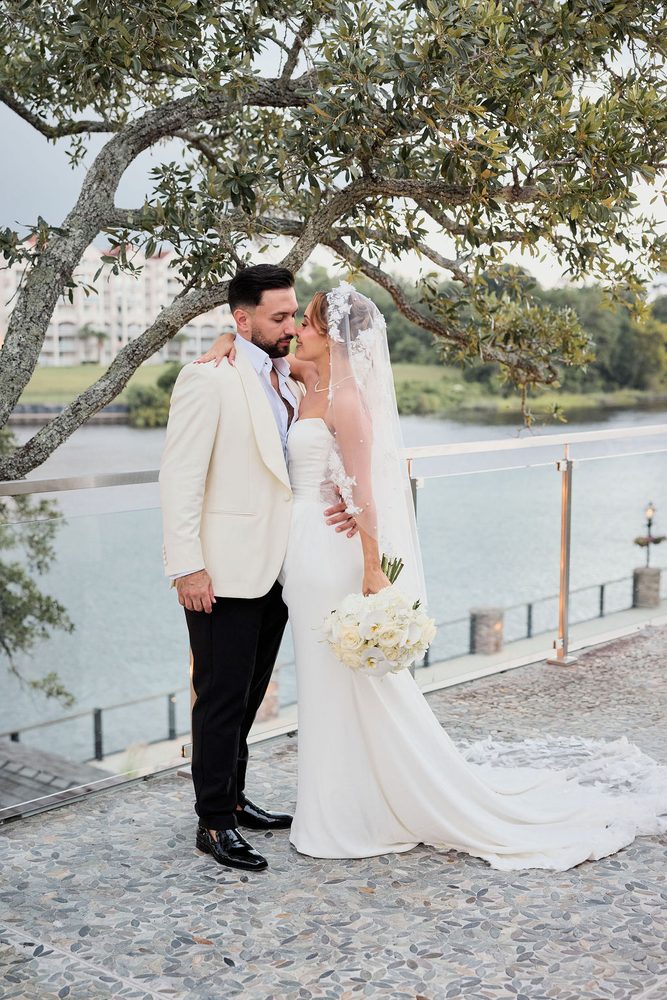 Couple leaning in nose to nose beneath oak branches, her long lace-edged train spread across the pebble terrace