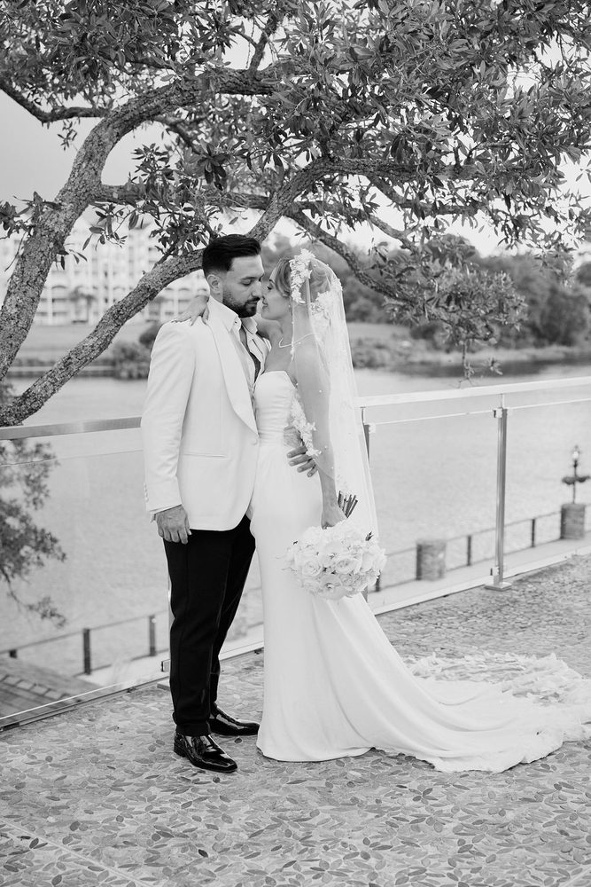Black-and-white portrait of bride and groom nose to nose under a sprawling tree beside a waterfront railing