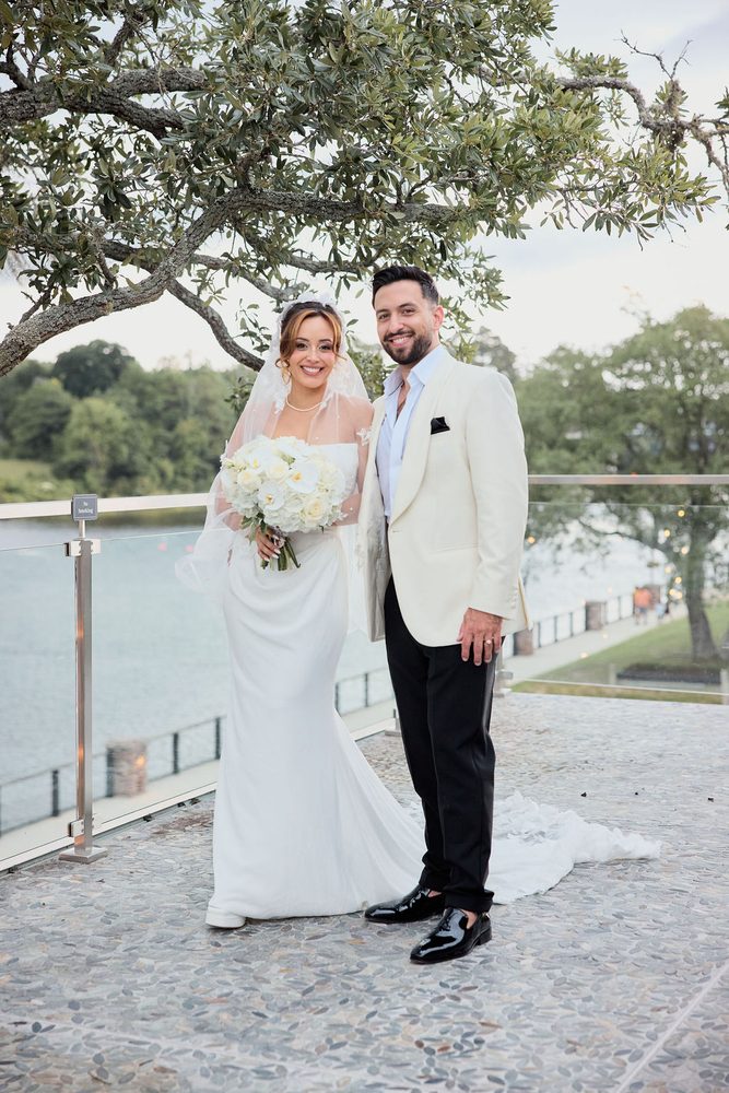 Bride and groom smiling at the camera on a riverside terrace, she holds white roses and wears a sheer veil