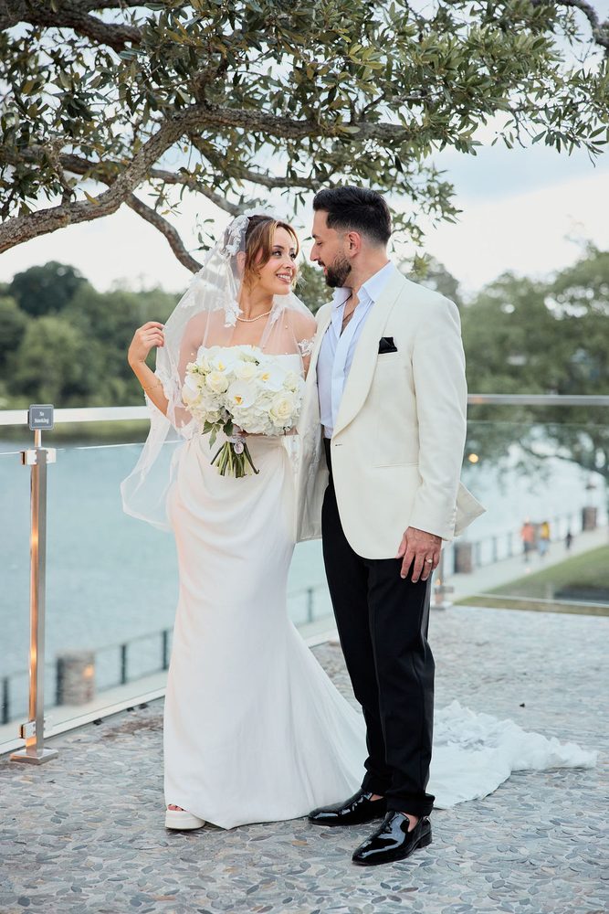Bride beaming at the camera while the groom gazes at her, white rose and orchid bouquet in hand, water behind