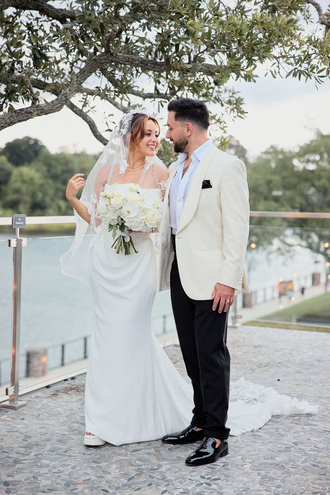 Bride touching her veil and smiling up at the groom in a cream dinner jacket on a lakeside terrace