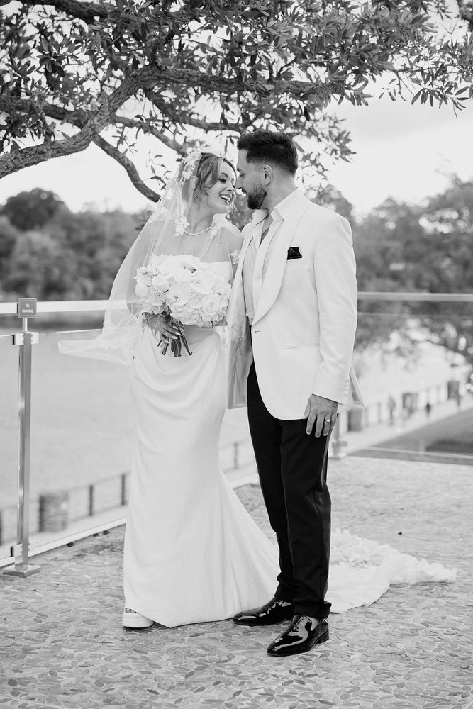 Black-and-white moment of the groom nuzzling a laughing bride, her veil blowing in the breeze by the railing