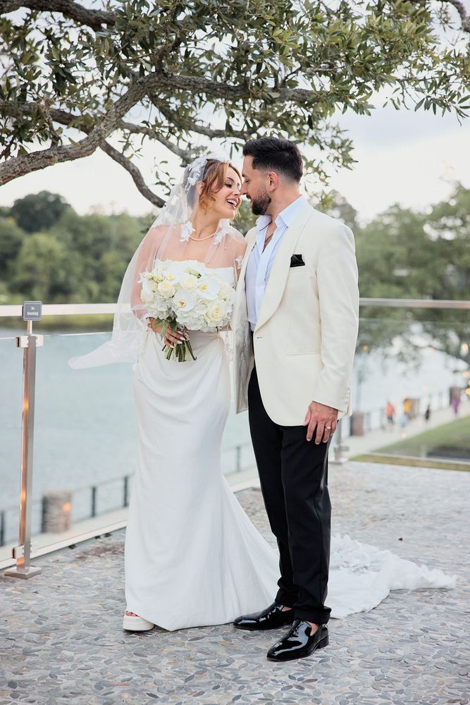 Groom leaning in to nuzzle the laughing bride, veil caught by the wind on a waterfront terrace at dusk