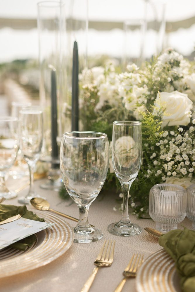 Reception place setting with gold-rimmed chargers, glassware, dark taper candles and a white rose centerpiece