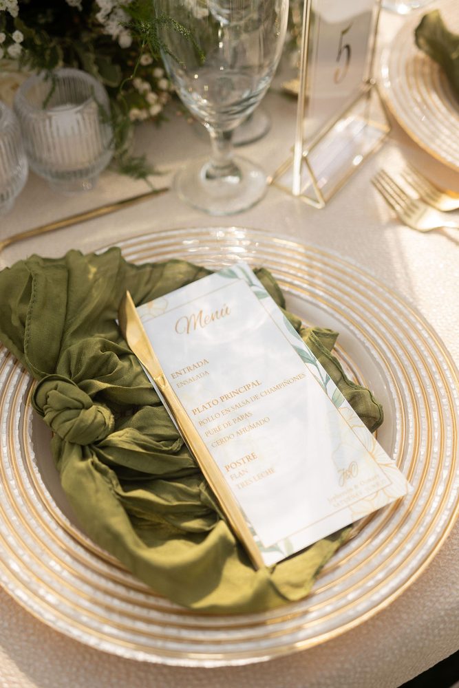 Gold-rimmed charger with a knotted olive-green napkin, gold knife and printed dinner menu beside a table number