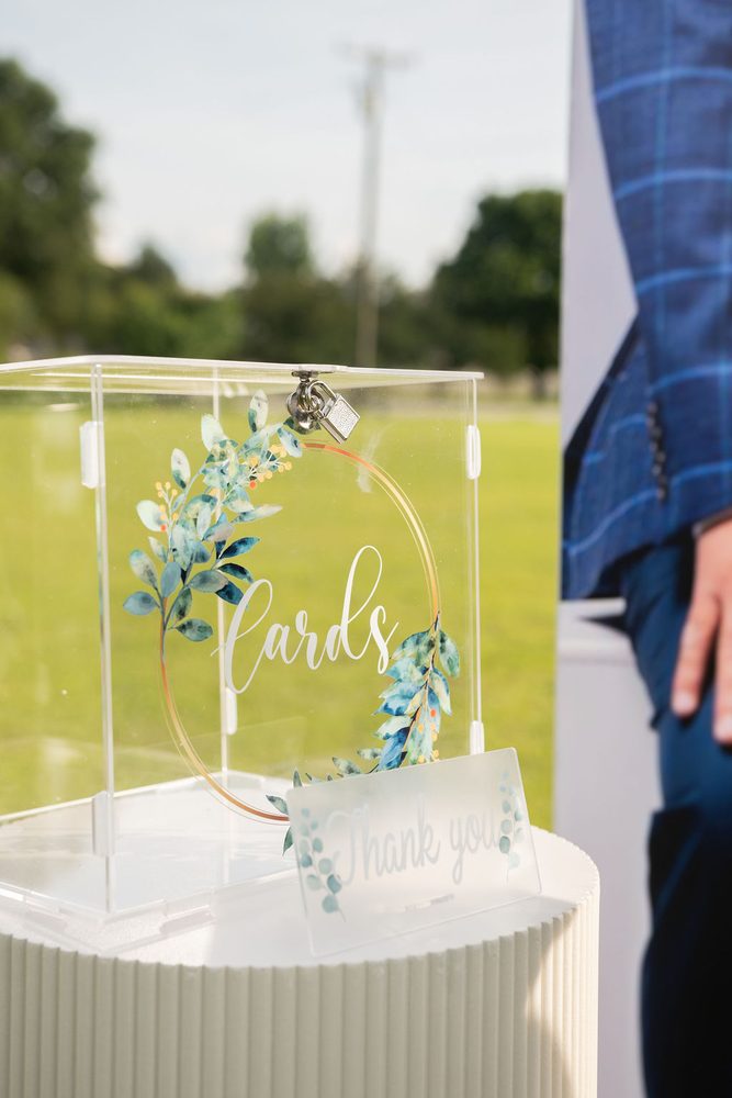 Clear acrylic card box with a eucalyptus wreath design and padlock on a white pedestal outdoors