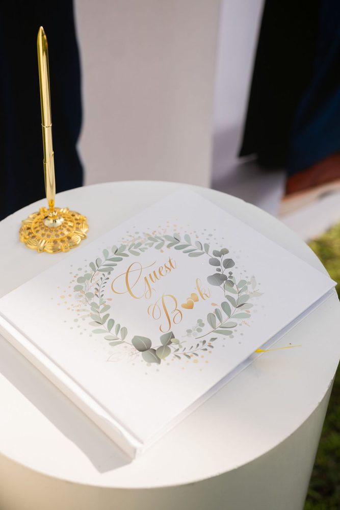 White wedding guest book with gold script and a eucalyptus wreath beside a gold pen stand
